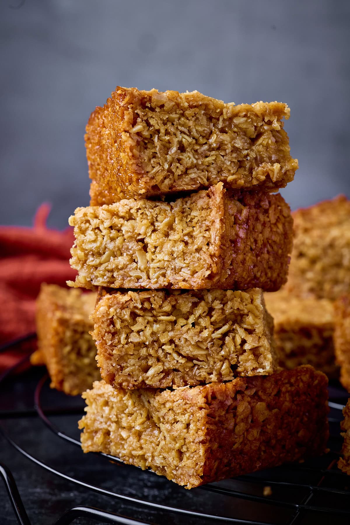 A tall shot of a stack of 4 flapjack squares, on top of a black wire cooling rack. In the left of the background, you can slightly see a red/ orange napkin, you can also see some more flapjacks stacked on top of each other in the background. This is all set on a black surface with a grey background.