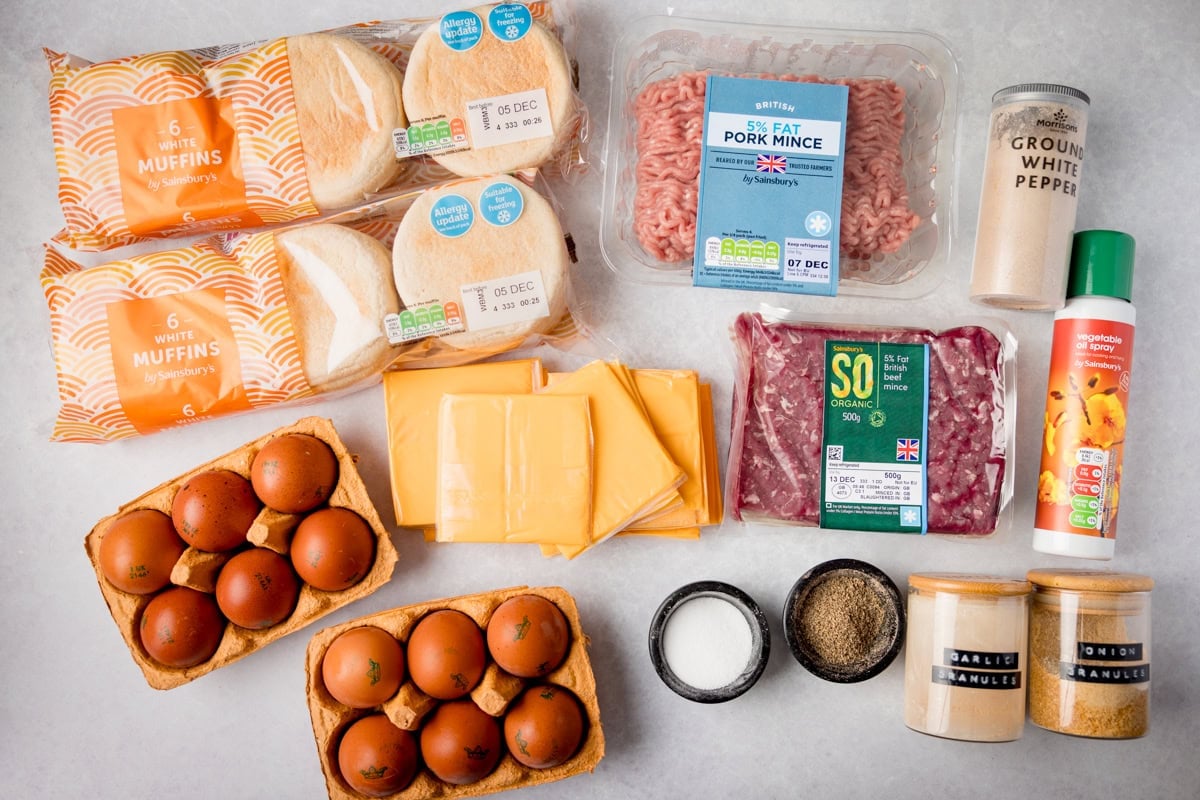 A wide, overhead shot of the ingredients for Homemade Sausage and Egg McMuffins. The ingredients are laid out on a white surface, they are as follows: minced pork, minced beef, salt, black pepper, white pepper, onion powder, garlic powder English bread muffins, and burger cheese.