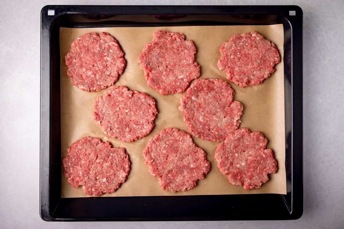 A wide, overhead prep shot of Sausage and Egg McMuffin. The image shows 8 raw patties laid out on some light brown parchment paper