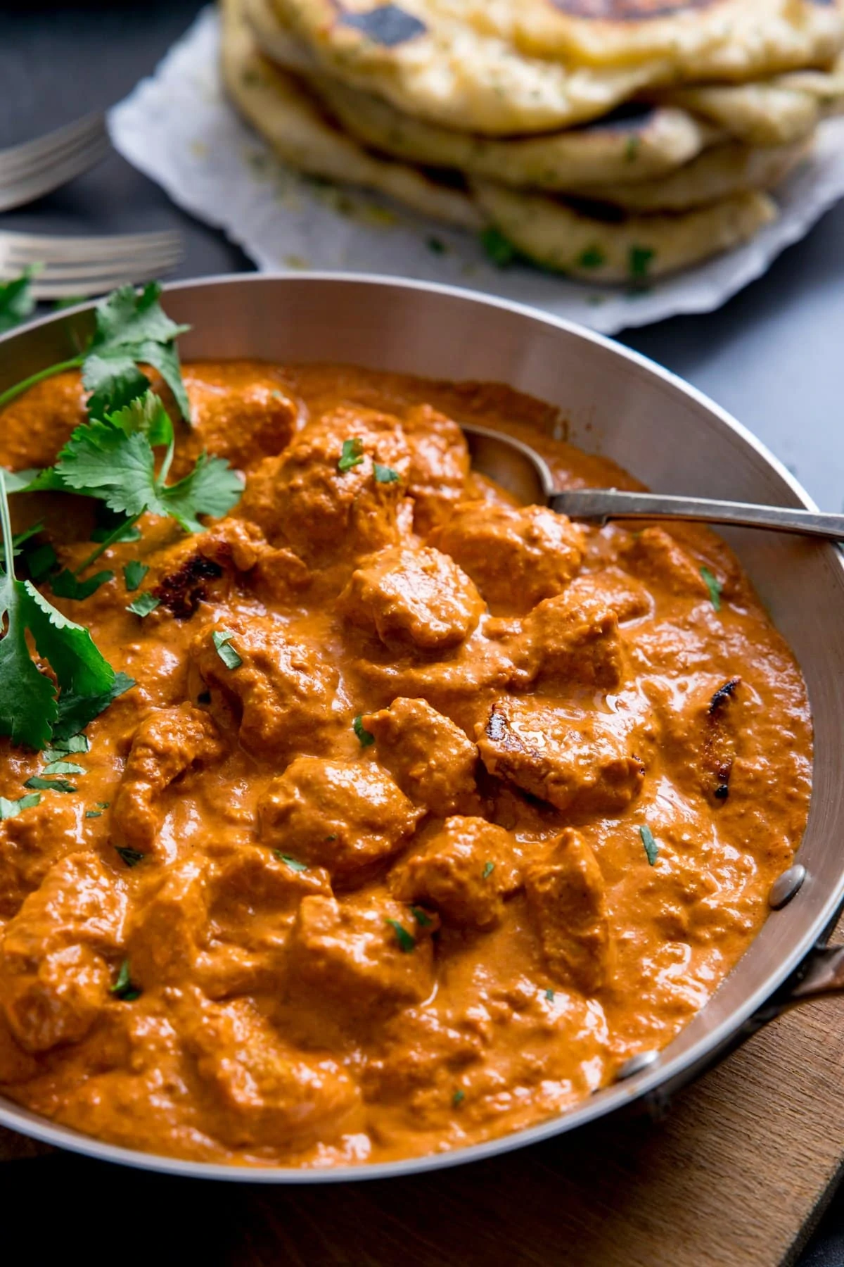 Silver frying pan with butter chicken curry in it. Pile of naan bread in the background.