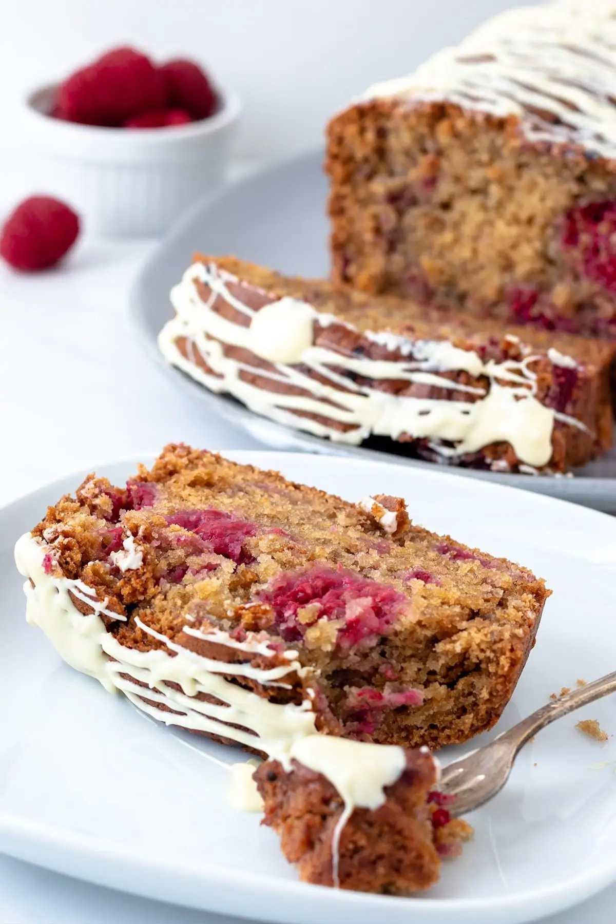 Close up image of a raspberry and white chocolate cake sat on a white plate showing the texture of the cake. There is the rest of the cake in the background out of focus.
